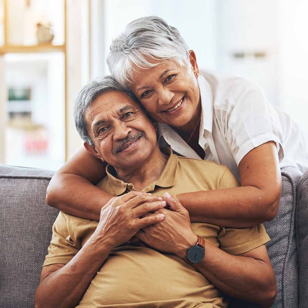 A senior couple smile and embrace on their couch.